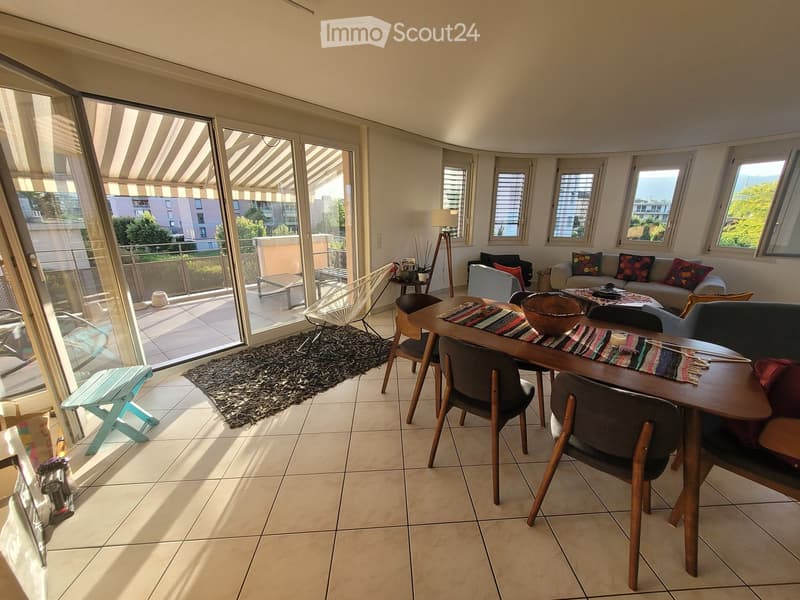 Bright living room with large windows and a balcony, featuring a sofa, dining table, chairs, and a rug.