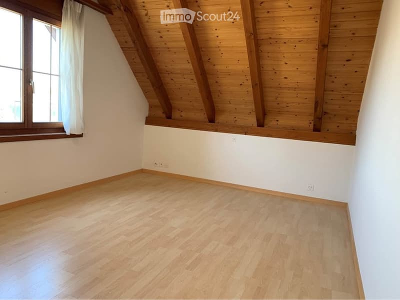 Empty room with wooden flooring, white walls, and exposed wooden beams on the ceiling. A window with white curtains is visible on one side.