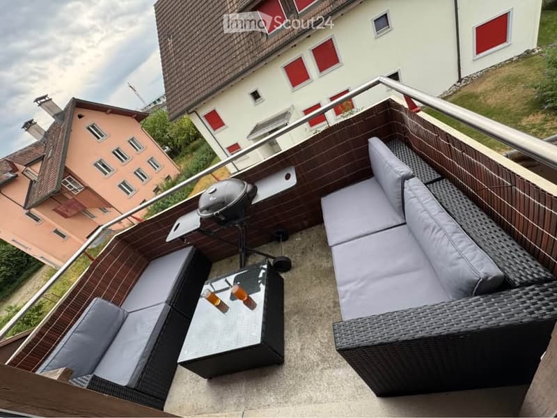 A modern rooftop terrace with a wicker sofa set, a glass coffee table, and a barbecue grill, overlooking a residential area with colorful houses.