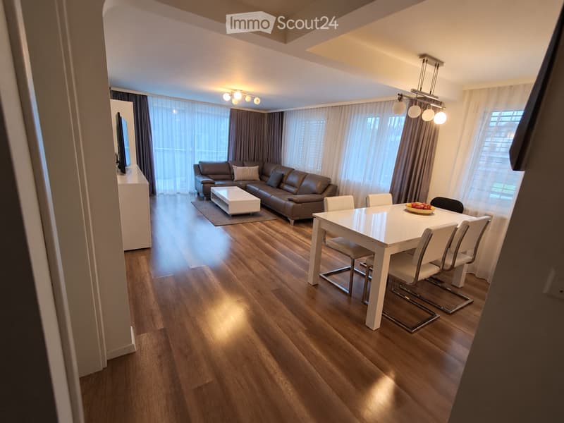 Modern living and dining area with wooden flooring, large windows with curtains, a brown leather sofa, a white coffee table, a white dining table with six chairs, and a TV on a white stand.