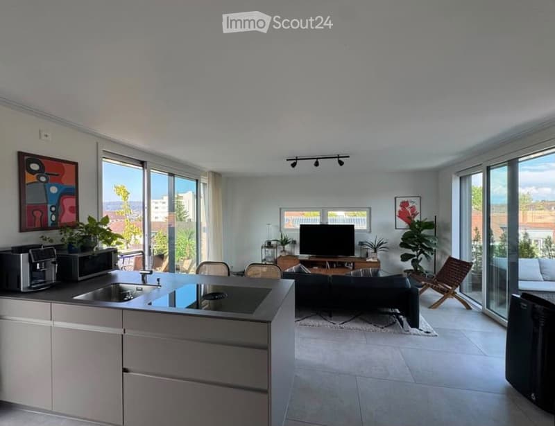 Modern open-plan living and kitchen area with large windows offering a view, featuring a black sofa, coffee table, TV, kitchen island with sink, microwave, and chairs.