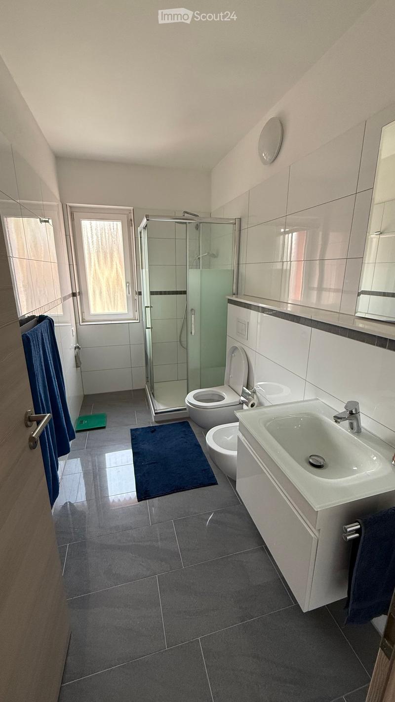 Modern bathroom with grey tiles, white fixtures, a glass-enclosed shower, a toilet, a sink, and a mirror. Blue towels are placed on the floor and a small green mat is near the shower.