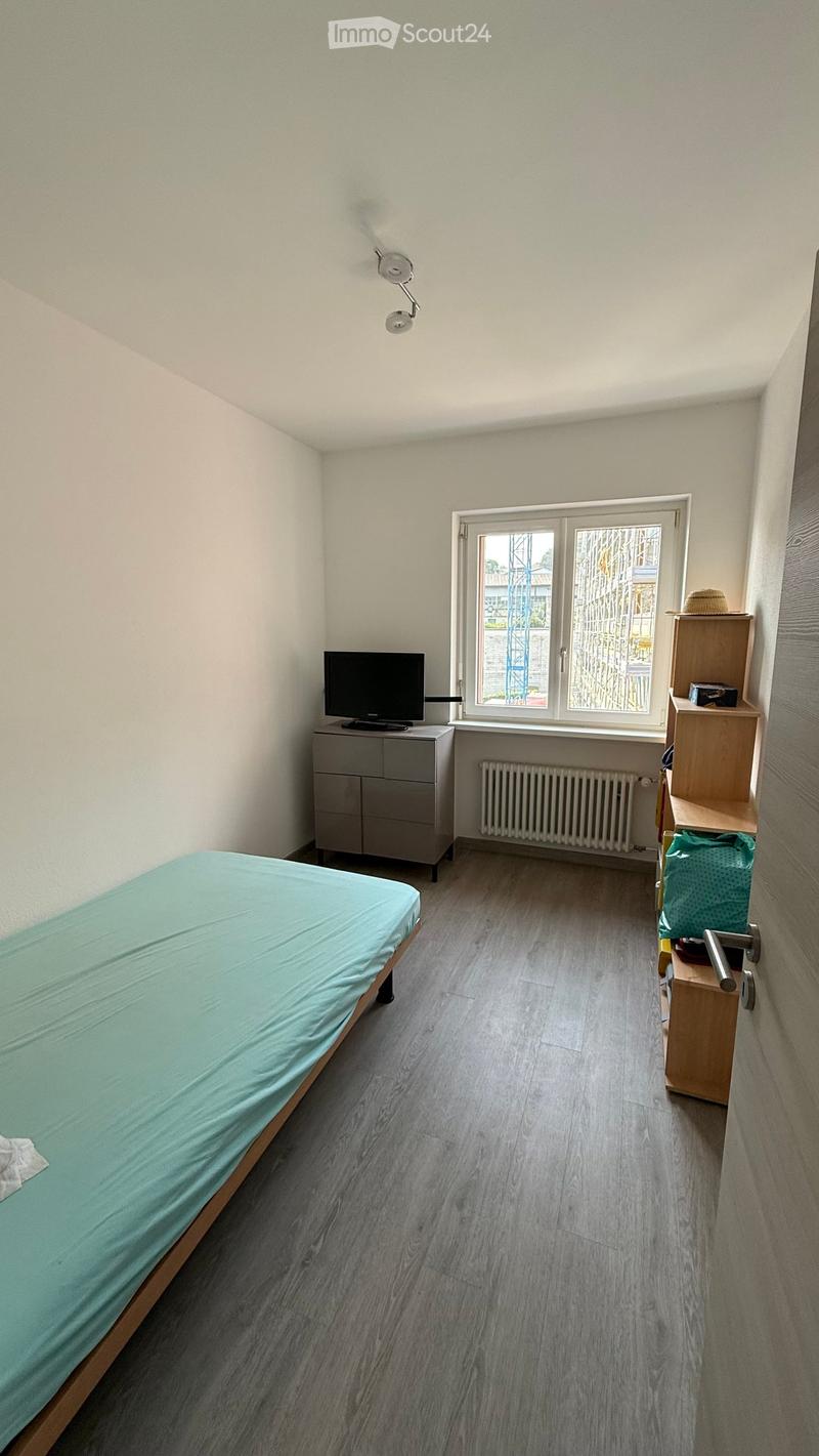 A small, furnished bedroom with a single bed, a television on a cabinet, a radiator, a shelf with a hat, and a window with a view of the outside.