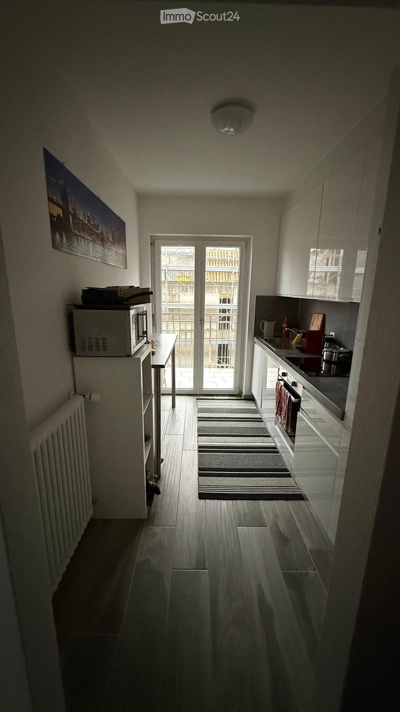 Modern kitchen with white cabinets, a microwave, oven, sink, and a small table near the entrance. The floor is wooden, and there is a striped rug in front of the door leading to a balcony.