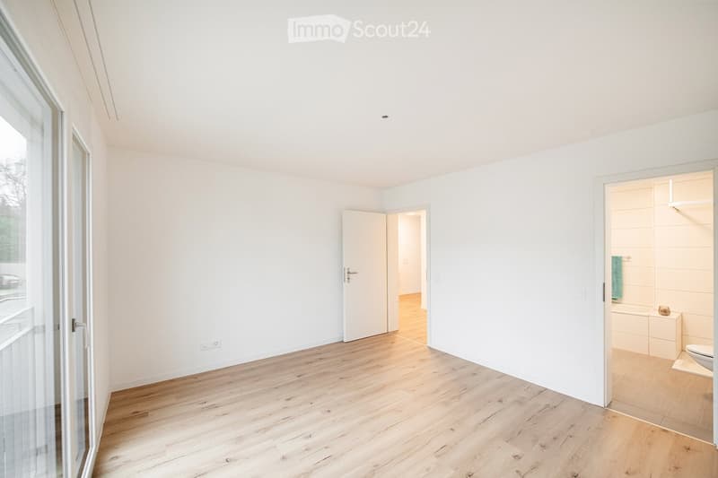 Empty room with light wooden flooring, white walls, and large windows. A partially visible bathroom with a toilet and a small shelf is seen through an open doorway.