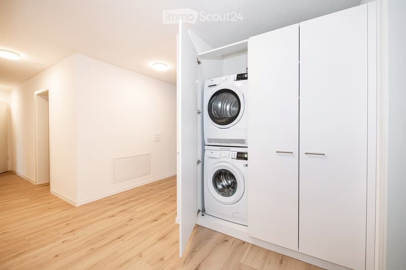 A small, clean, unfurnished room with light wooden flooring and white walls, featuring a built-in closet with a private washing machine and dryer unit.