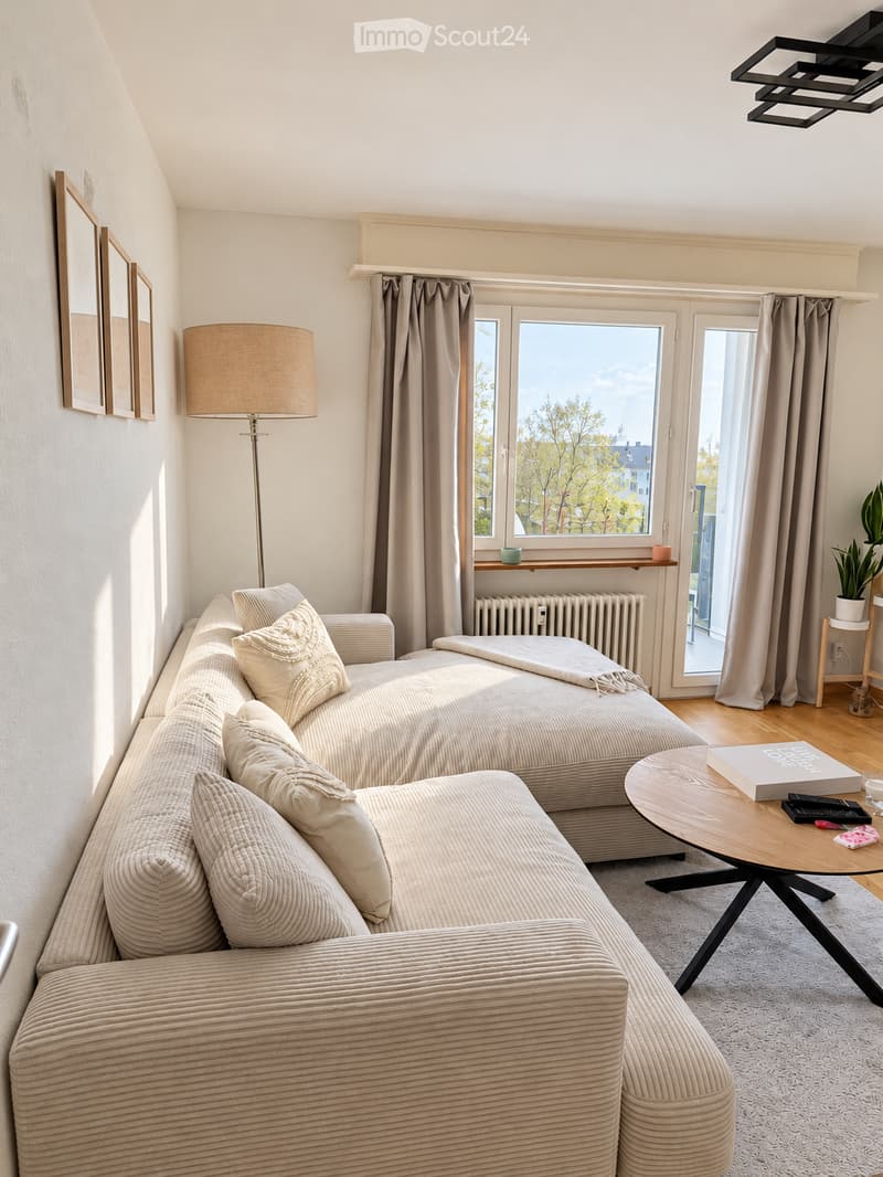 Modern living room with a sofa, coffee table, floor lamp, and large windows offering a view of trees and buildings.