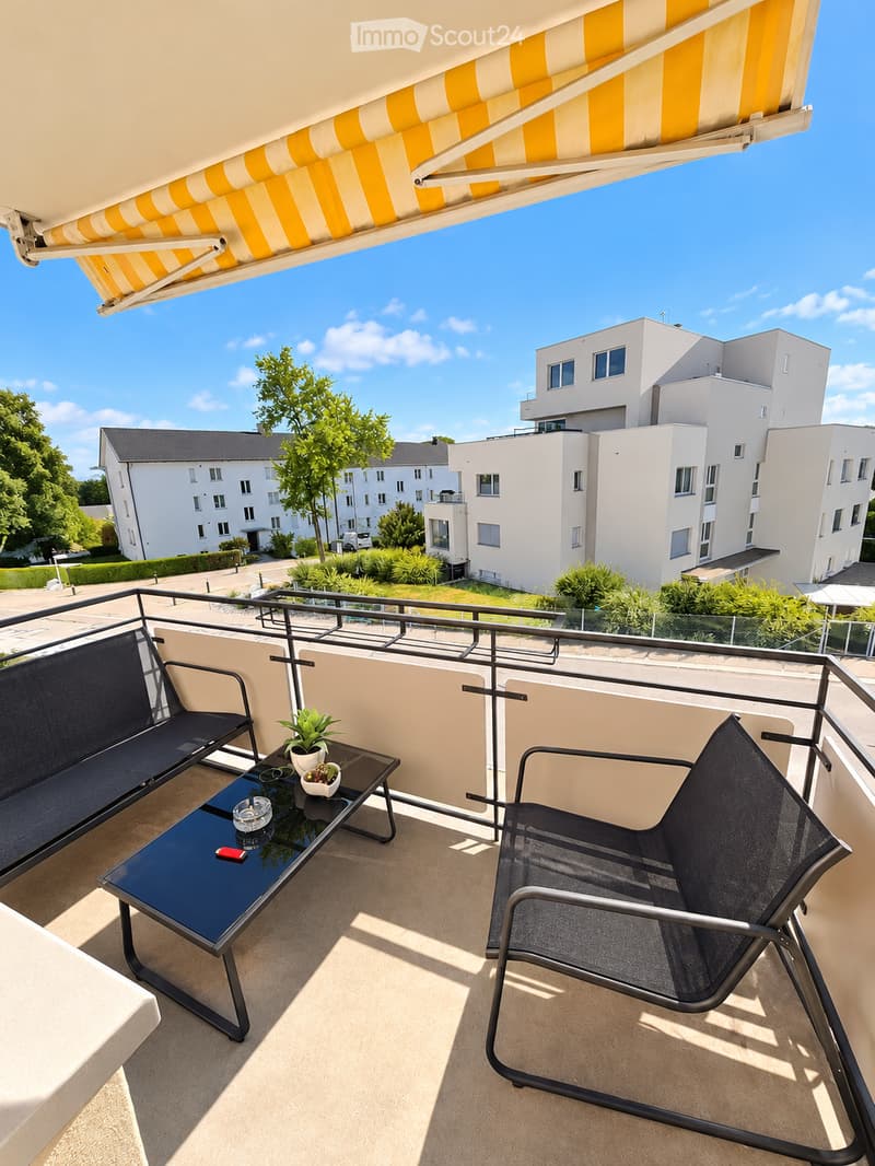 Balcony with a black sofa, a black table, a black chair, and a small potted plant. The balcony has a metal railing and is covered by a yellow and white striped awning.