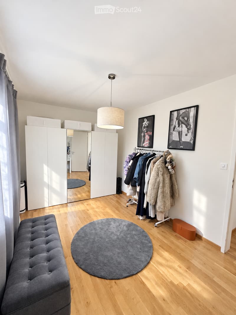 Bright, modern living space with wooden flooring, a grey tufted sofa, a round grey rug, a large white wardrobe with mirrored doors, a coat rack with jackets, and two framed pictures on the wall.