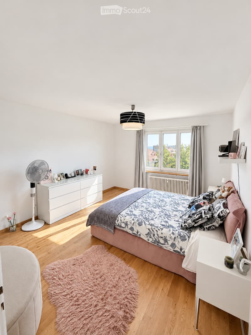 Bright bedroom with wooden flooring, large window with curtains, bed with patterned bedding, nightstand, fan, dresser, and decorative items.