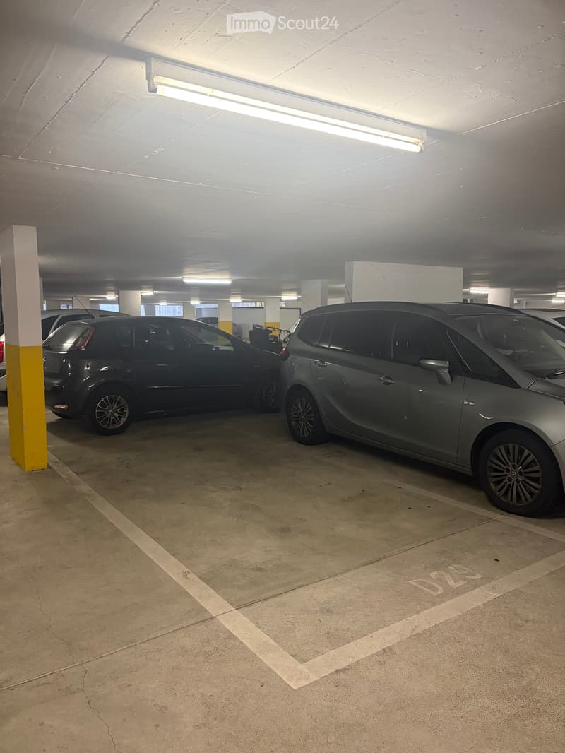 Underground parking area with two parked cars, concrete floor, fluorescent lighting, and support pillars.