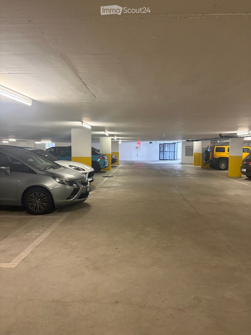Underground parking area with several parked cars, concrete pillars, and fluorescent lighting.
