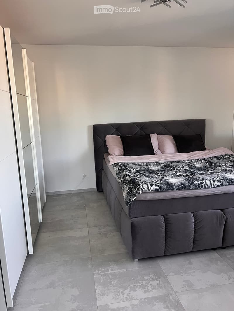 Modern bedroom with a large bed featuring a tufted headboard, multiple pillows, and a fur throw. The room has light grey walls and a concrete floor.