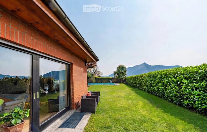 Well-maintained garden with a lawn, neatly trimmed hedges, outdoor seating, and a swimming pool in the background, with a view of mountains.