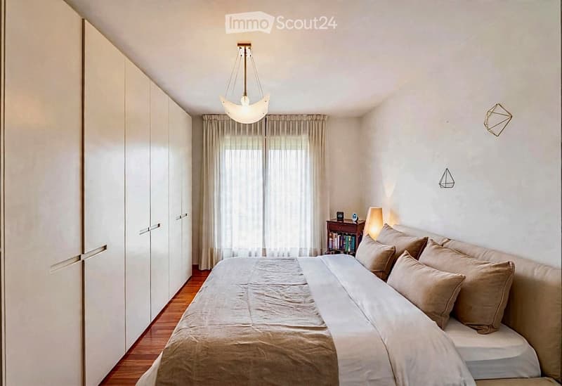 Modern bedroom with a large bed, nightstand, books, pendant light, and built-in wardrobe.