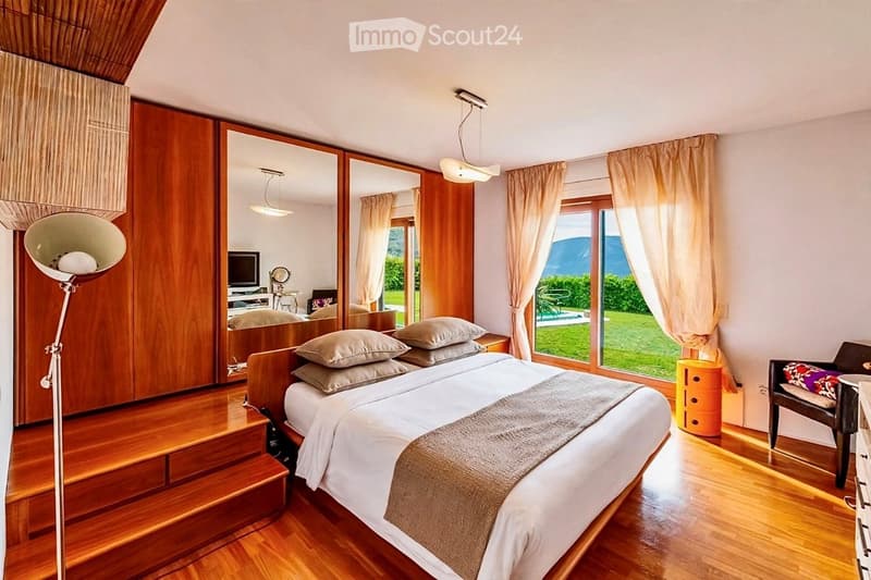 Elegant bedroom with a large bed, multiple pillows, a wooden floor, large windows with curtains, a floor lamp, and a mirrored wardrobe.