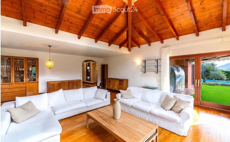 Bright living room with exposed wooden beams, white sectional sofa, coffee table, decorative vase, wooden cabinets, and large glass doors leading to a garden.