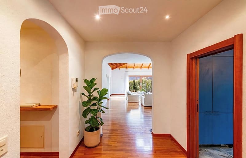 Elegant hallway with wooden flooring, arched doorways, and a potted plant, leading to a bright living area with white furniture and a view of greenery outside.
