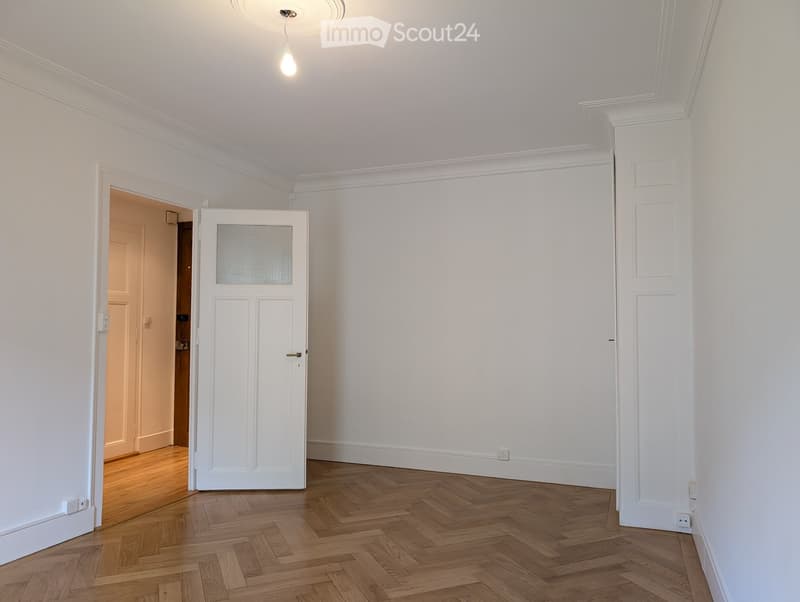 Empty room with white walls, wooden herringbone flooring, and a white door with frosted glass panel. The room has a ceiling light fixture and visible electrical outlets.