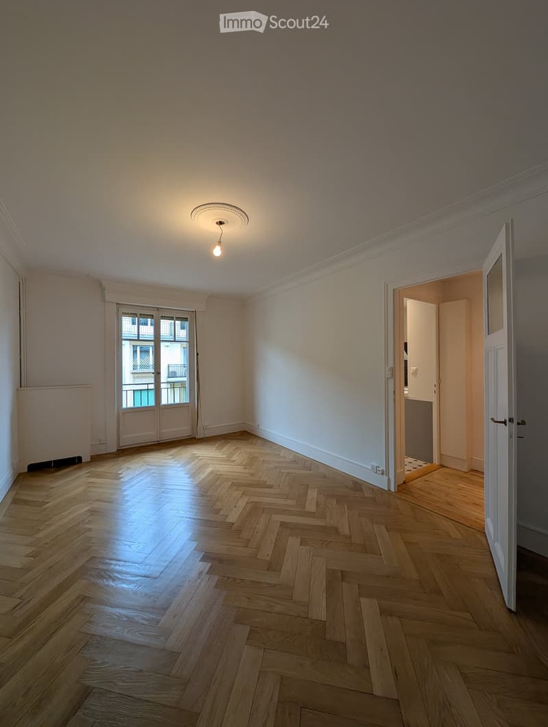 Empty room with wooden herringbone flooring, white walls, and a ceiling light fixture. Features a large window with a view of a balcony and an open doorway leading to another room.