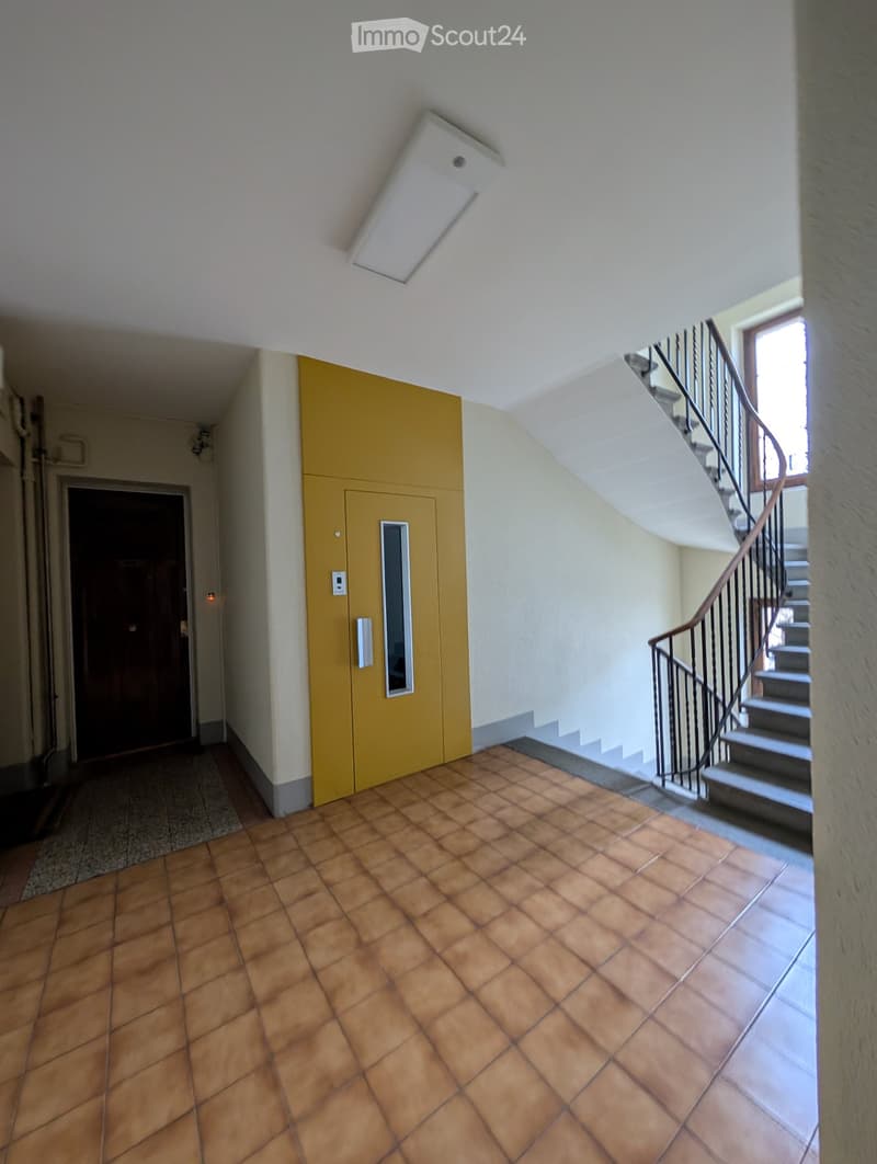 Hallway with tiled floor, staircase with metal railing, yellow elevator, and recessed ceiling light.