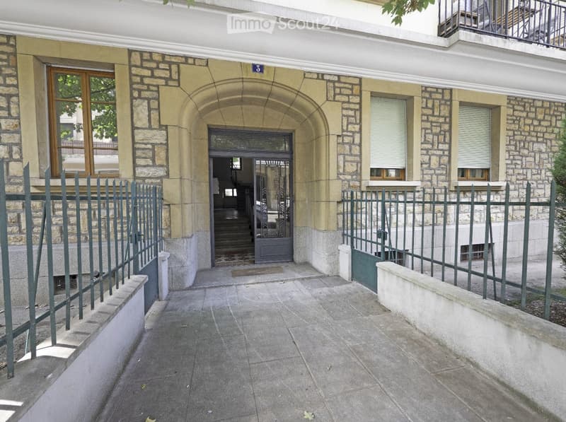 Entrance to a multi-story building with stone facade, arched doorway, and metal gate. The entrance features a tiled floor and a small step leading up to the door.