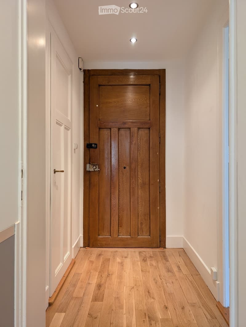 A narrow hallway with light wooden flooring, white walls, and a large wooden door at the end. The hallway has recessed ceiling lights and a small window on the right side.