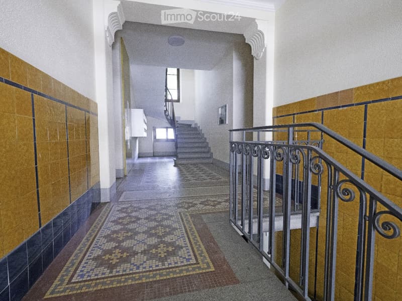 A long, well-lit hallway with patterned mosaic tiles, yellow and blue wall tiles, and a metal staircase leading upwards.