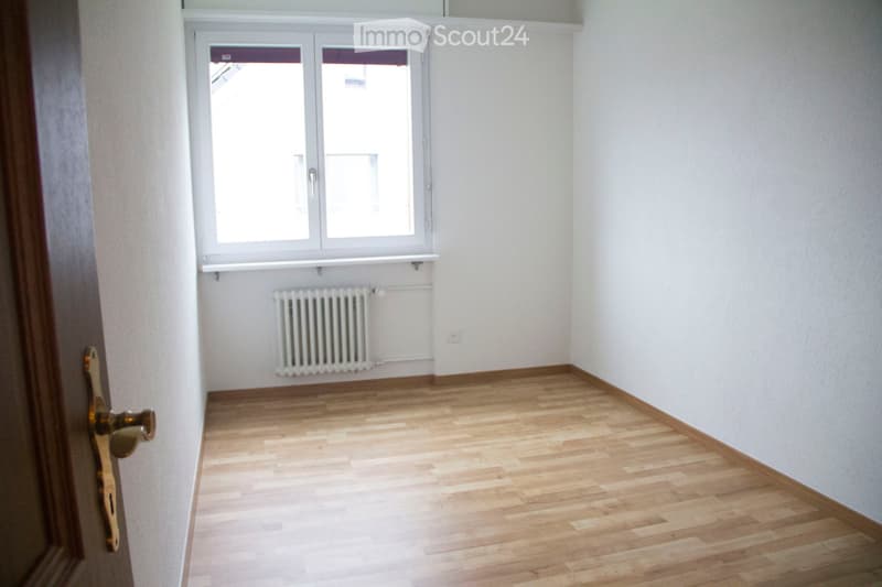 Empty room with wooden flooring, white walls, a window with blinds, a radiator, and a door with a brass handle.