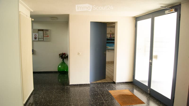 A clean, well-lit hallway with tiled flooring, white walls, and a green vase with flowers placed near a bulletin board. A door leads to another room, and a glass door is visible on the right side.