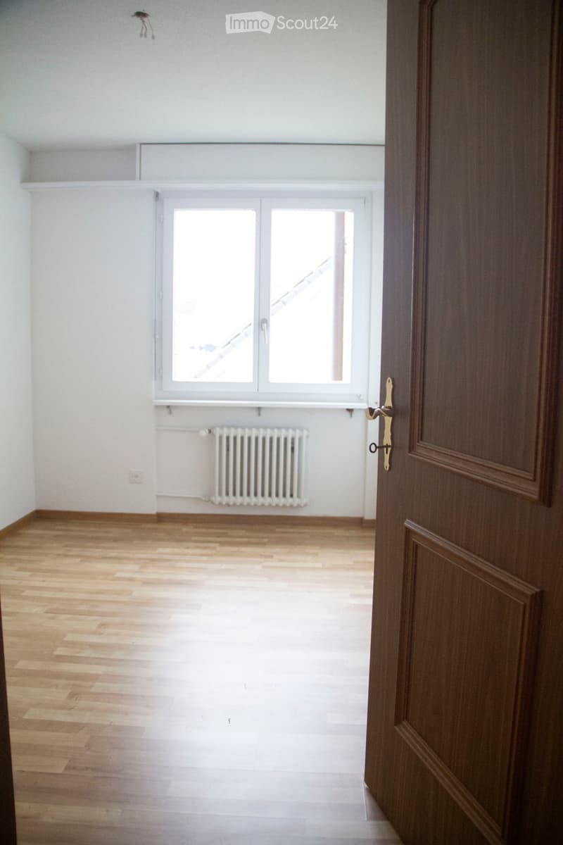 Empty room with wooden flooring, white walls, a window with white frame, and a radiator below the window. A wooden door is partially visible on the right side.