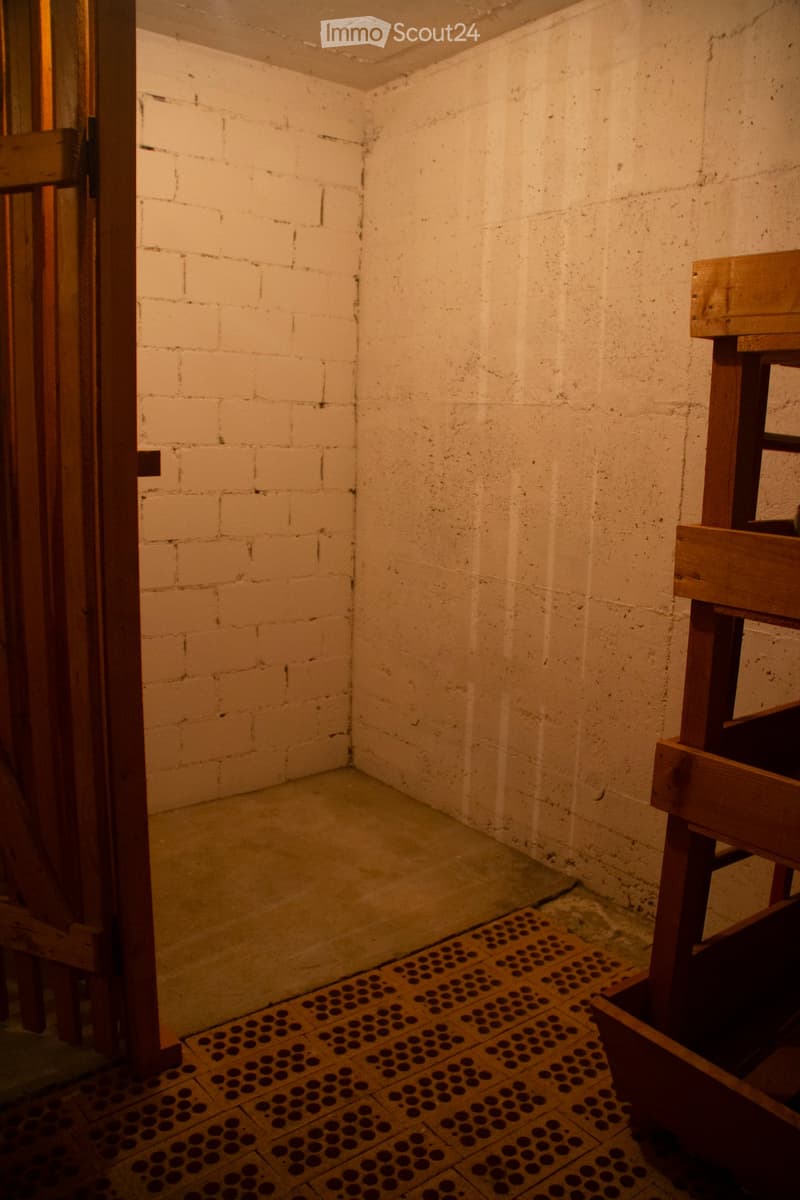 Empty room with exposed brick walls, concrete floor, and wooden door frame. A wooden stool is partially visible on the right side.