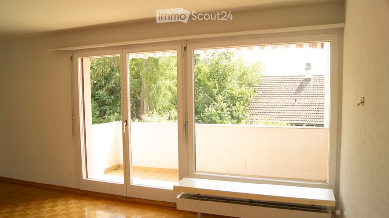 Empty room with wooden flooring, large sliding glass doors leading to a balcony, white walls, and a radiator under the window.