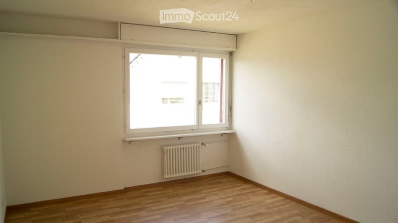 Empty room with wooden flooring, white walls, a window with white frame, and a radiator below the window.