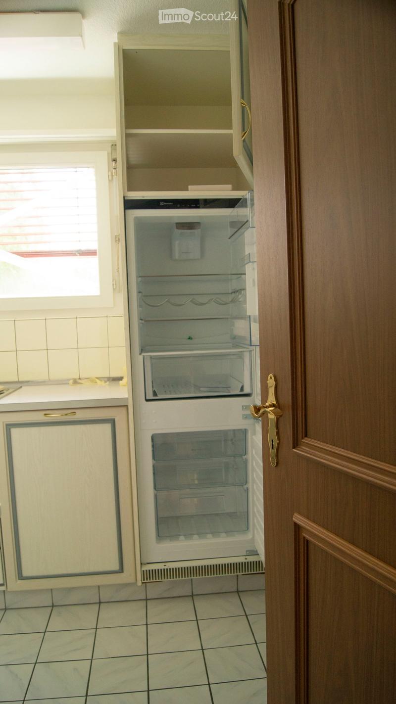 A small kitchen area with a white refrigerator and light-colored cabinets, featuring tiled flooring and a window with blinds.