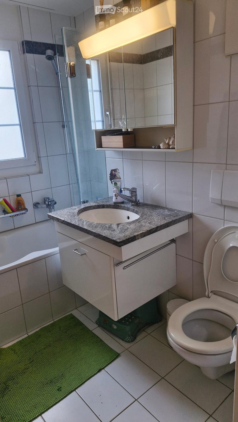 Modern bathroom with a white tiled floor, granite countertop sink, toilet, bathtub, and a large mirror with built-in storage. The space includes a green bath mat and a glass shower enclosure.