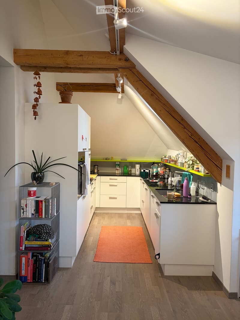 Modern kitchen with white cabinets, wooden beams, and a black countertop. Features include a sink, refrigerator, and various kitchen items on the counter.
