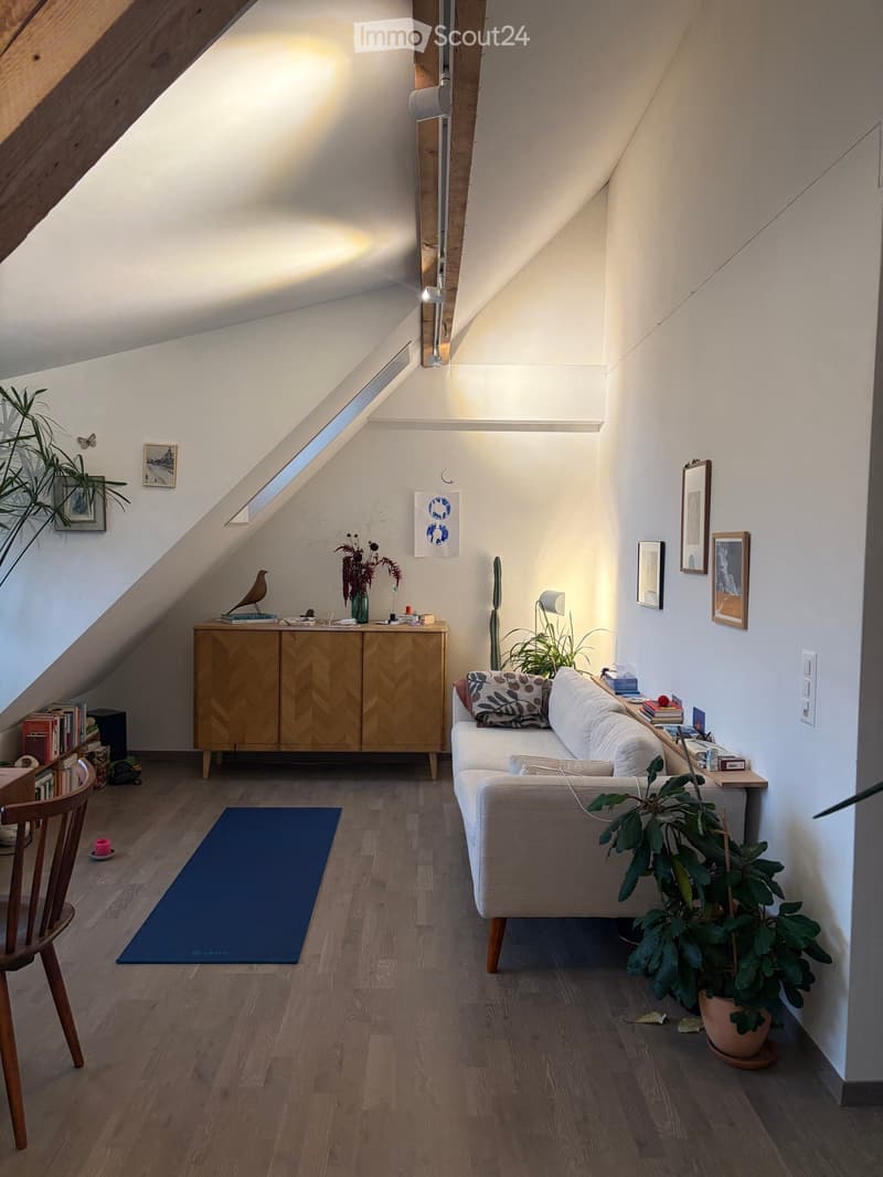 Cozy attic living room with wooden floors, slanted ceilings, and large windows. Features a sofa, wooden cabinet, chair, small table, and several plants.