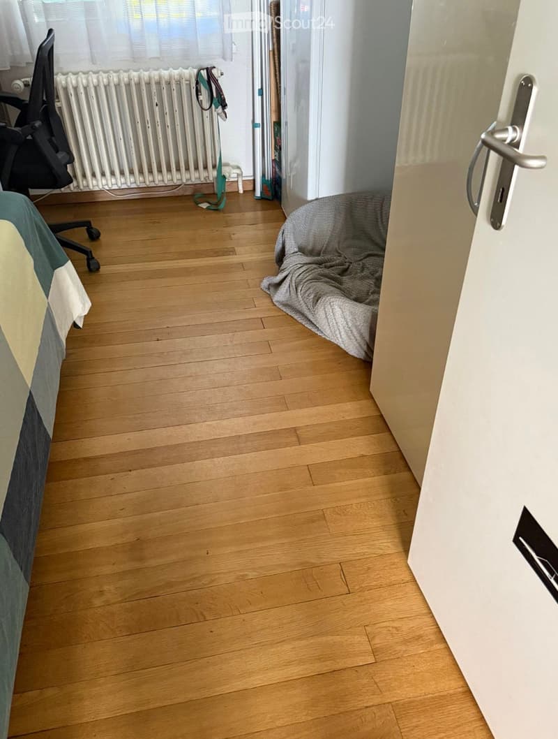 Empty room with wooden flooring, a radiator, and a partially visible bed and chair in the background.