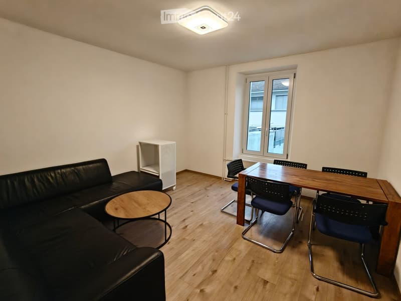 A modern, well-lit room with a black leather sofa, a small round wooden coffee table, a wooden dining table with four chairs, and a white shelving unit.