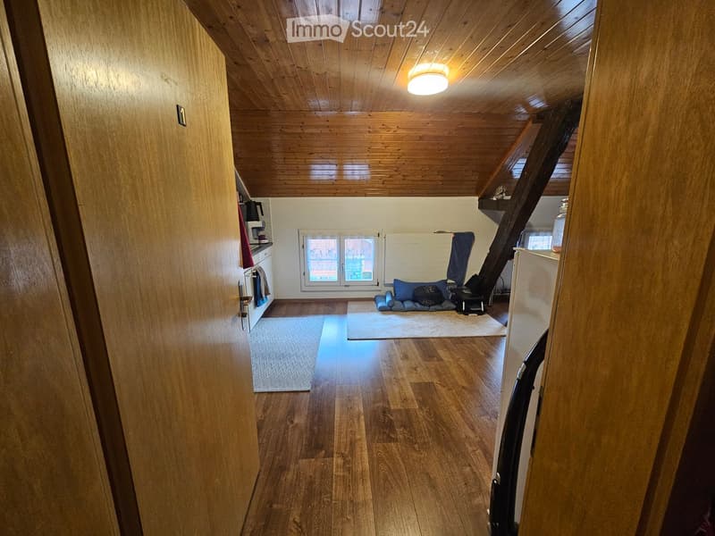 A cozy, well-lit room with wooden floors and a slanted wooden ceiling. The space includes a small seating area with a sofa and a table, a kitchen area with cabinets and appliances, and a window letting in natural light.