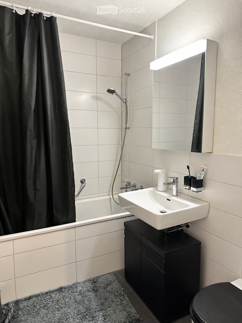 Modern bathroom with a bathtub, sink, mirror, and toilet. Features a black shower curtain, a grey bath mat, and a sleek black cabinet under the sink.