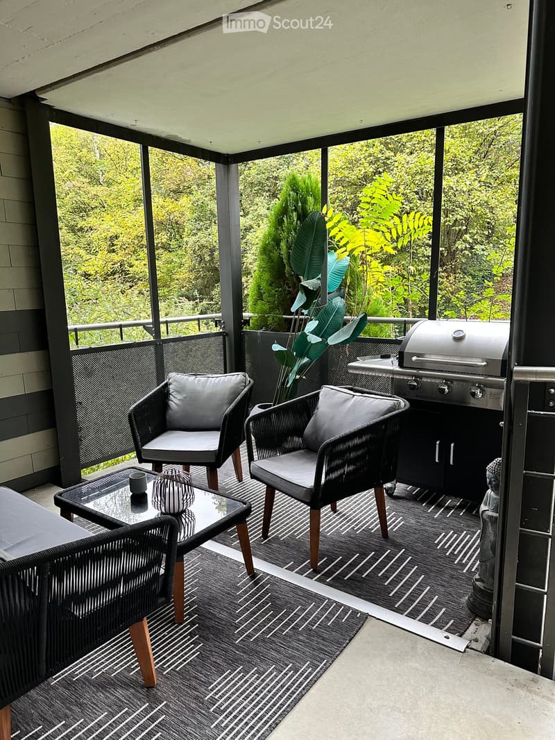 A modern, enclosed patio with wicker chairs, a glass coffee table, a grill, and lush greenery visible through the glass walls.