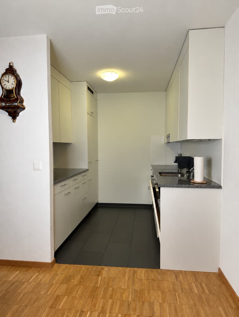 Modern kitchen with white cabinets, black countertops, and a dark tiled backsplash. Features a sink, faucet, oven, and a wall-mounted clock.
