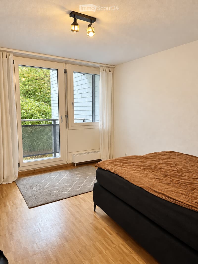 Bedroom with wooden flooring, a bed with a brown blanket, a grey rug, and large windows with curtains leading to a balcony.