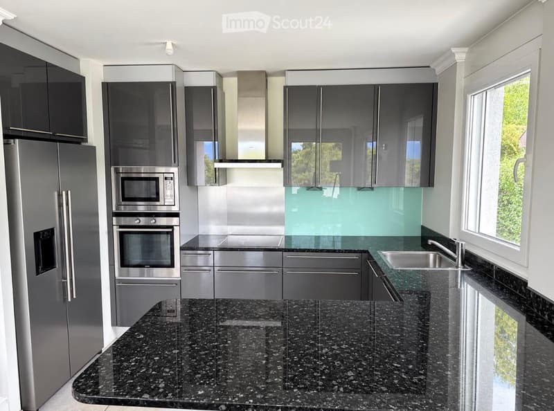 Modern kitchen with dark grey cabinets, stainless steel appliances, granite countertops, and a large window offering a view of greenery.