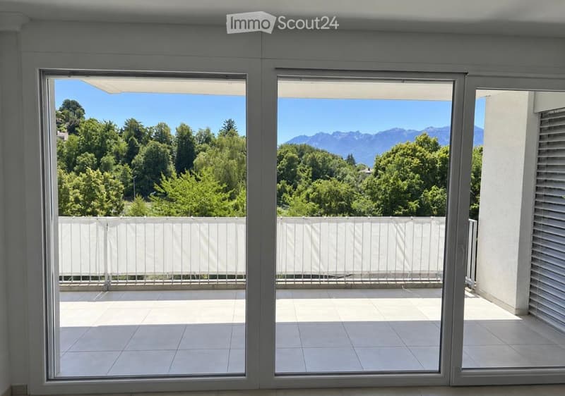 Large, modern balcony with white tiled flooring, white metal railing, and expansive views of green trees and distant mountains.