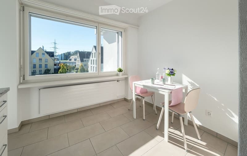 Bright room with tiled floor, white walls, large windows, a small table with chairs, and a vase with flowers on the table.