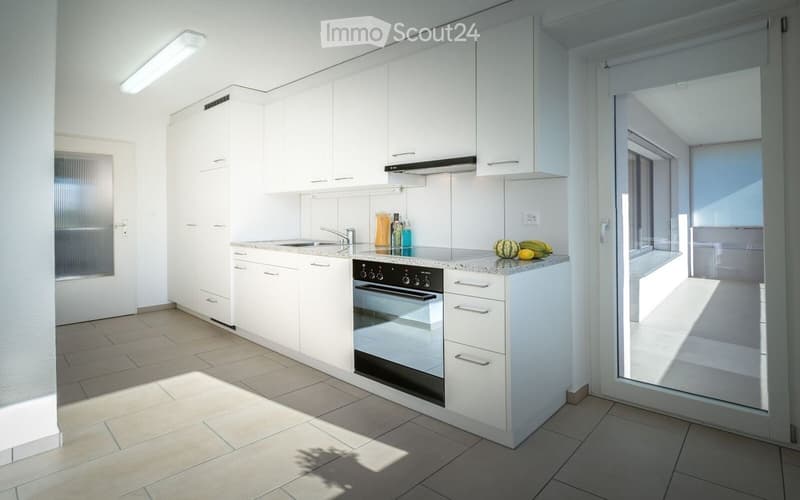 Modern kitchen with white cabinets, granite countertop, built-in oven, and sink. The space is bright with natural light coming through large windows and a sliding glass door leading to a balcony.
