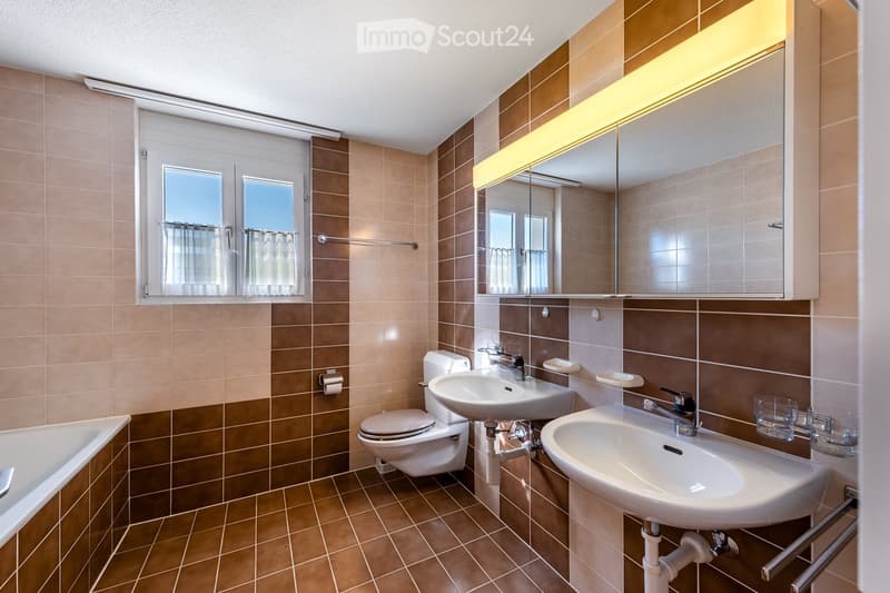 Modern bathroom with beige and brown tiles, a bathtub, toilet, two sinks, a large mirror with built-in lighting, and a window with curtains.